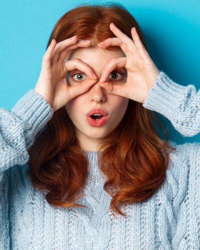 Funny redhead female model in sweater, staring at camera through fingers glasses, seeing something interesting, standing over blue background.