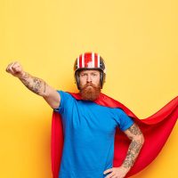 Self confident serious superhero man has superhuman power, makes flying gesture, ready for flight and helping people, saves world, wears headgear, red cloak and blue t shirt, has brave behaviour